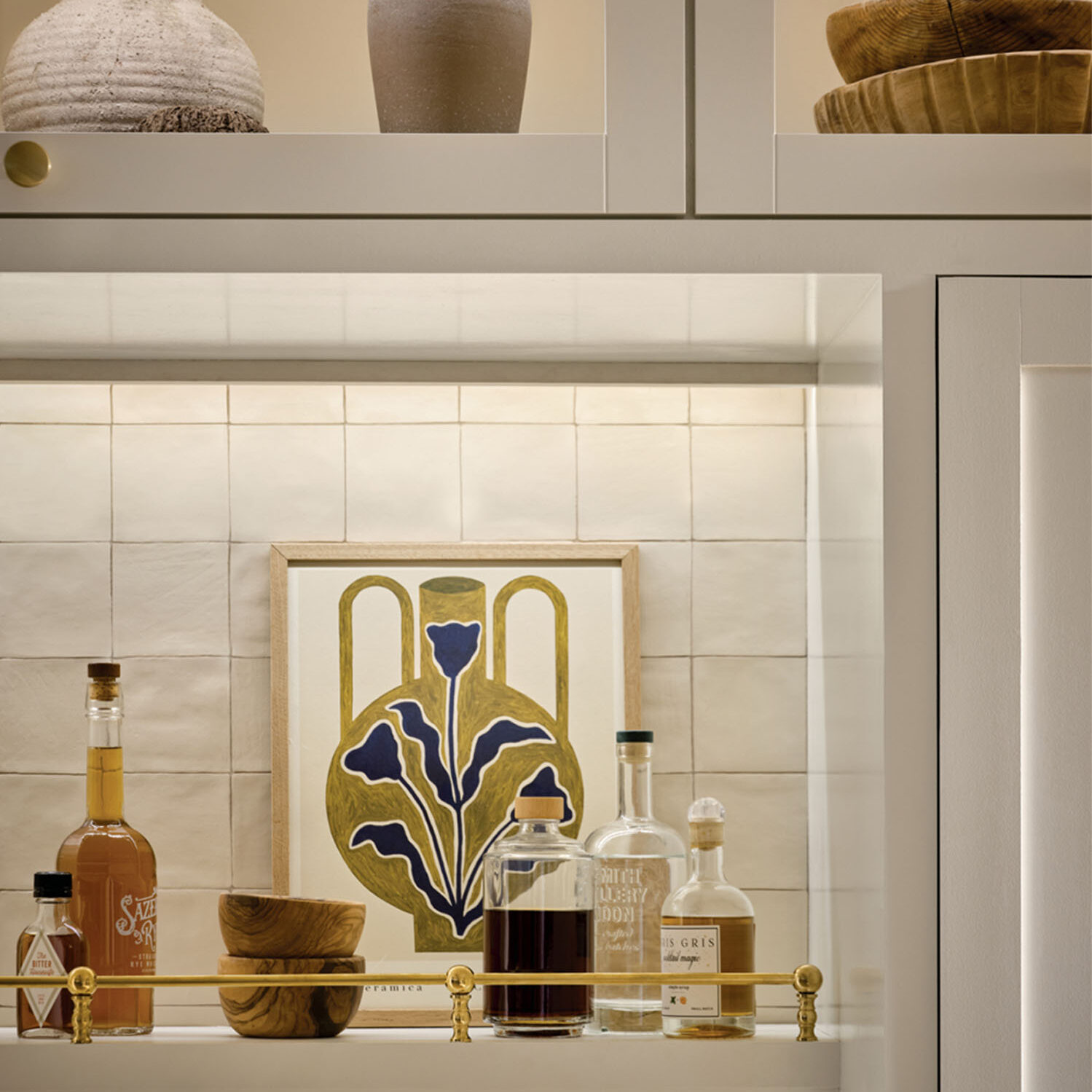 Decorative white shelving with undercabinet lighting, with bottles, wood bowls, and floral art on a tiled backsplash.