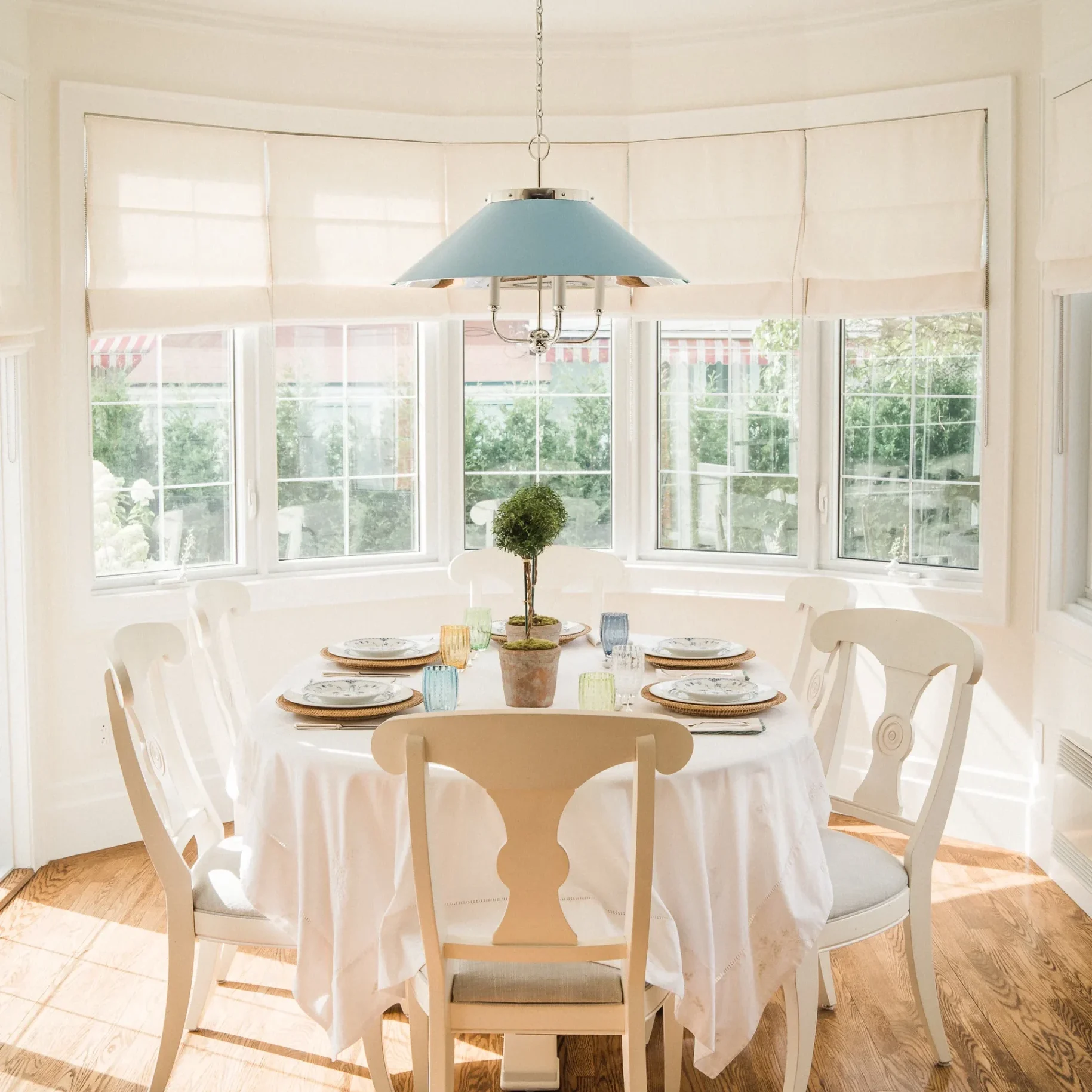 Casual dining nook with a light wood pedestal table set for four, surrounded by large windows, and a light blue pendant light.