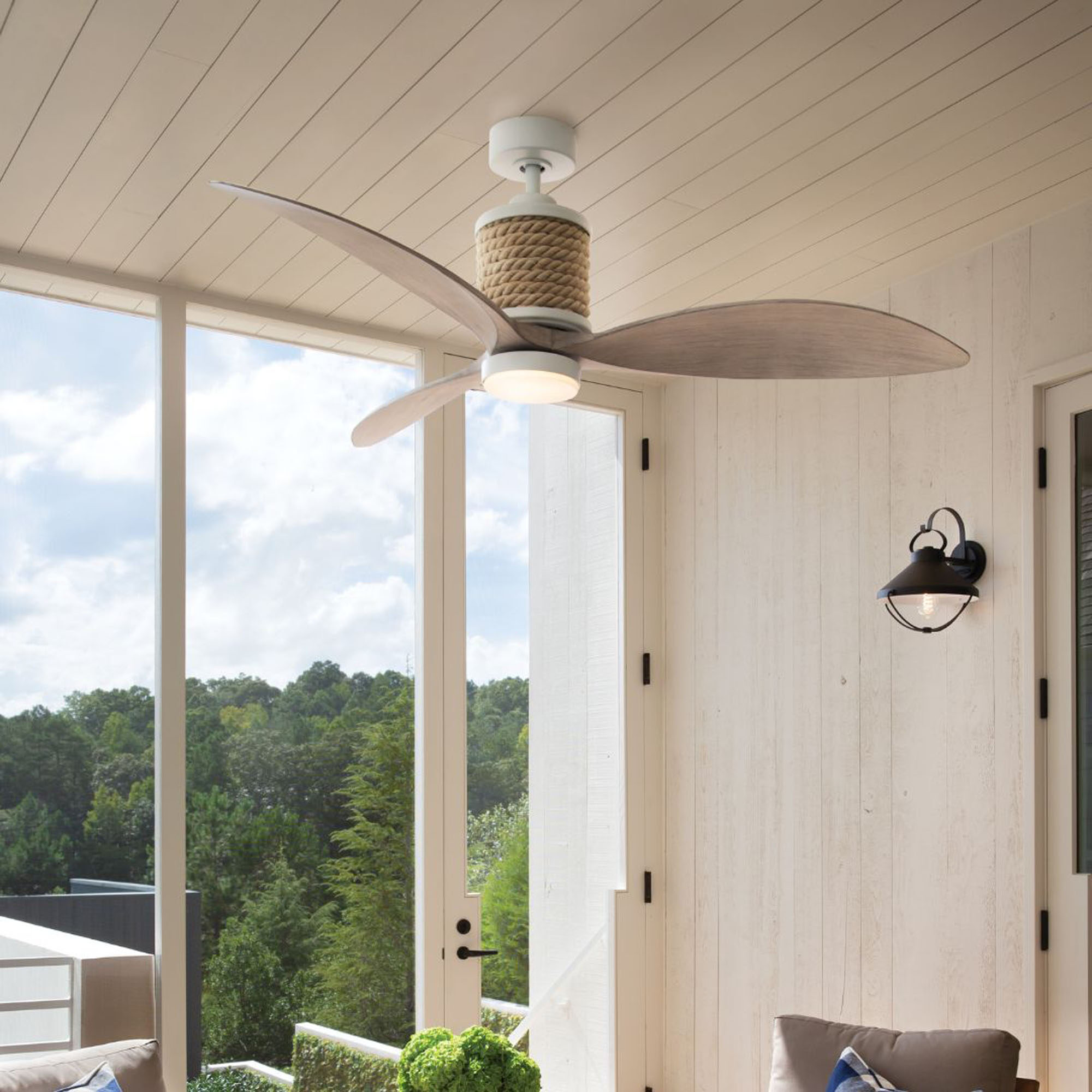 Rustic white-paneled room with a large window view of a lush forest, featuring a bronze ceiling fan.