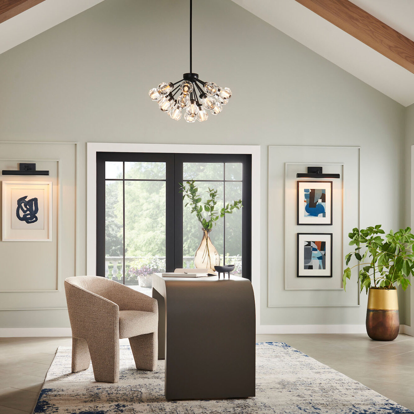 Home office with high vaulted ceilings and exposed wood beams, featuring a modern Sputnik-style chandelier. A large, curved desk sits in the center, flanked by a sculptural beige chair, two pieces of framed art lit by picture-frame sconces, and a large black window.
